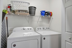 Ein Waschraum mit zwei weißen Whirlpool-Waschmaschinen, einem Feuerlöscher, einem grünen Kehrblech, einem weißen Wäschekorb, einem blauen Eimer und einem Regal mit diversen Gegenständen.