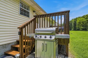 Ein tragbarer Gasgrill mit vier Brennern, einer Bedienblende und einem Schornstein, platziert auf einer Holzterrasse vor einem Haus.