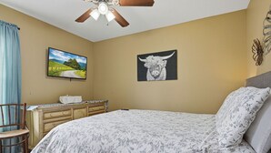 Een slaapkamer met een bed, een bureau, een stoel, een plafondventilator, een televisie en twee kunstwerken aan de muur.