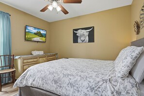 Ein Schlafzimmer mit einem Bett, einem Schreibtisch, einem Sessel, einem Deckenventilator, einem Fernseher und zwei Wandbildern.