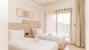 Ein Hotelzimmer mit zwei Betten, einem großen Fenster mit Vorhängen und drei gerahmten Bildern an der Wand.