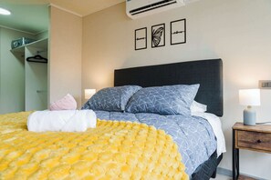 Ein ordentlich bezogenes Bett mit gemusterten Betttextilien, ein Nachttisch mit Lampe und Wandkunst.