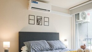 Un dormitorio con cama, mesita de noche, lámpara, ventana con vistas y aire acondicionado.