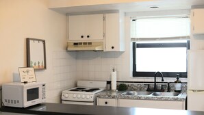 Une cuisine avec des meubles blancs, une cuisinière, un four à micro-ondes, un évier et une fenêtre avec des stores.