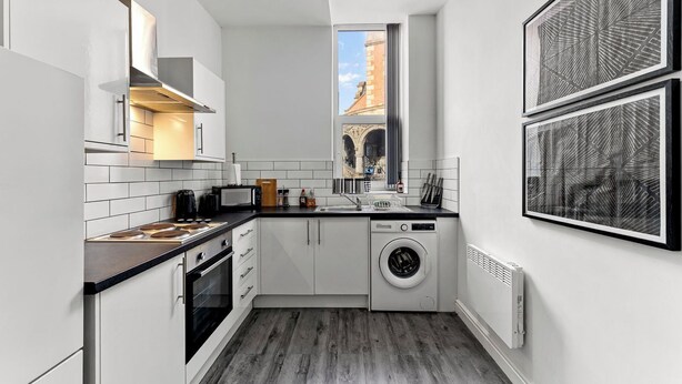A compact kitchen with a built-in oven, microwave, and washing machine, a window with a view of a building, and a framed artwork on the wall. - Modern 1BR Flat in Leeds - WiFi (Leeds)