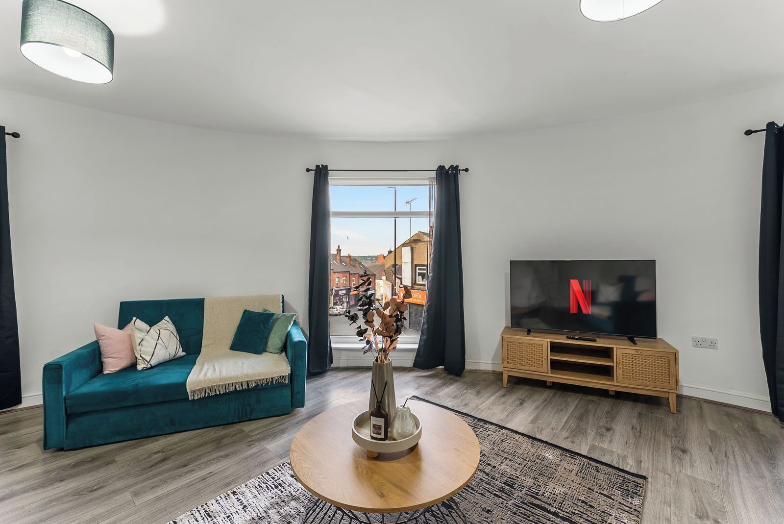 Un salon avec un canapé de couleur turquoise, une table basse ronde en bois, un téléviseur posé sur un meuble en bois et une vue sur la rue par une fenêtre.