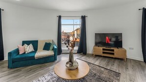 Ett vardagsrum med en turkosfärgad soffa, ett runt soffbord i trä, en tv på ett träställ och utsikt över gatan genom ett fönster.