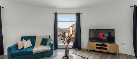 A living room with a teal sofa, a round wooden coffee table, a TV on a wooden stand, and a view of the street through a window.