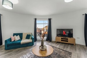 Ein Wohnzimmer mit einem blaugrünen Sofa, einem runden Holztisch, einem Fernseher auf einem Holzgestell und Blick auf die Straße durch ein Fenster.