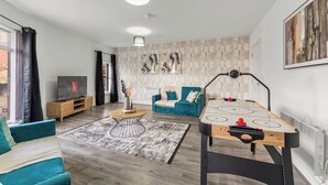 A modern living room with a flat-screen TV, a blue sofa, a coffee table, and a shuffleboard table.