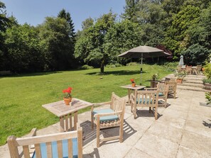 Eine Terrasse im Freien mit Holzmöbeln, einem Tisch und Stühlen, umgeben von einer Rasenfläche und Bäumen.