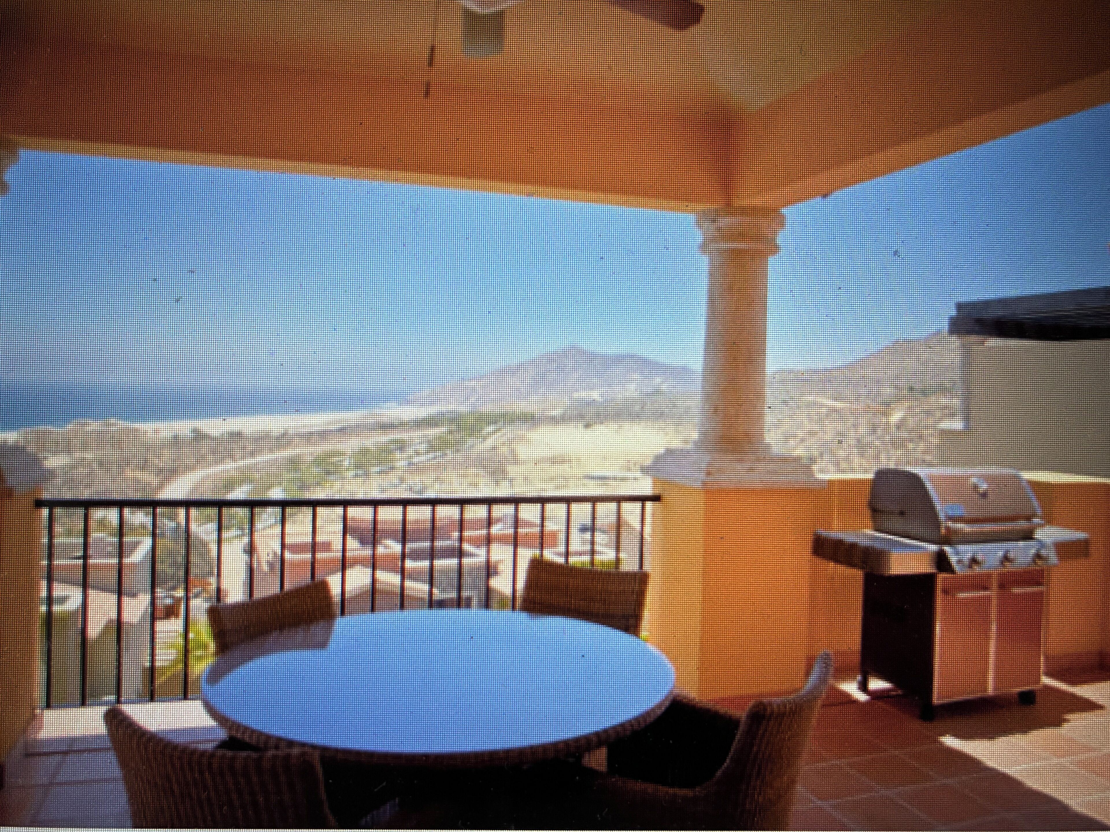 A balcony with a round table and chairs, a grill, and a view of a desert landscape.
