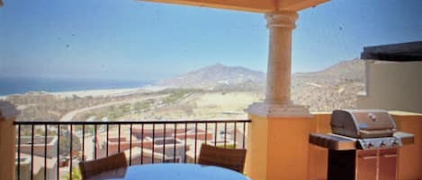 A balcony with a round table and chairs, a grill, and a view of a desert landscape.