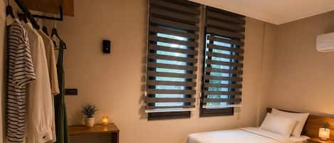A bedroom with two beds, a small stool, a nightstand with a lamp, a wall-mounted air conditioner, and a window with blinds.