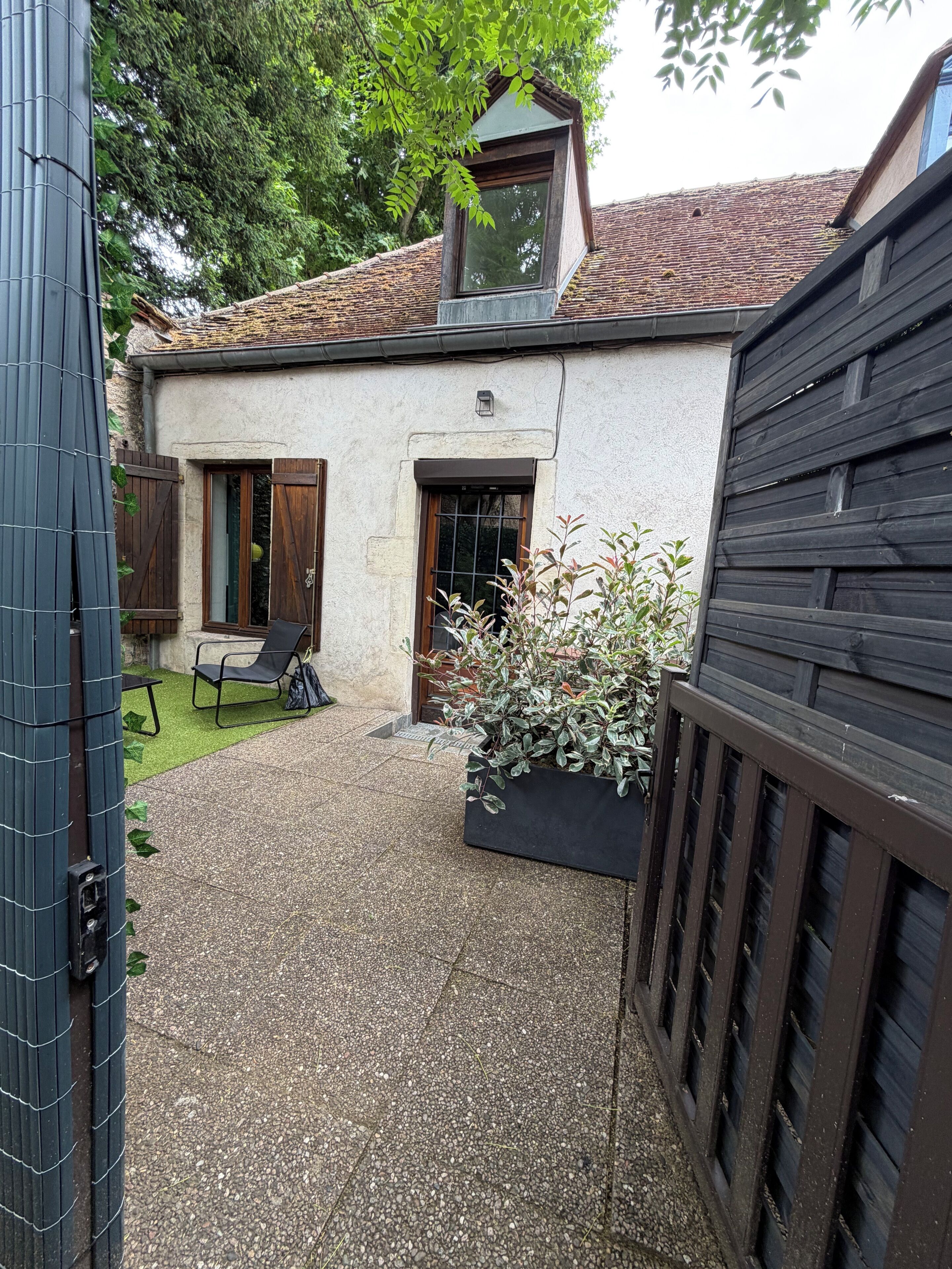 Un bâtiment avec un toit en tuiles, un petit jardin agrémenté d’un banc et un portail en métal noir.