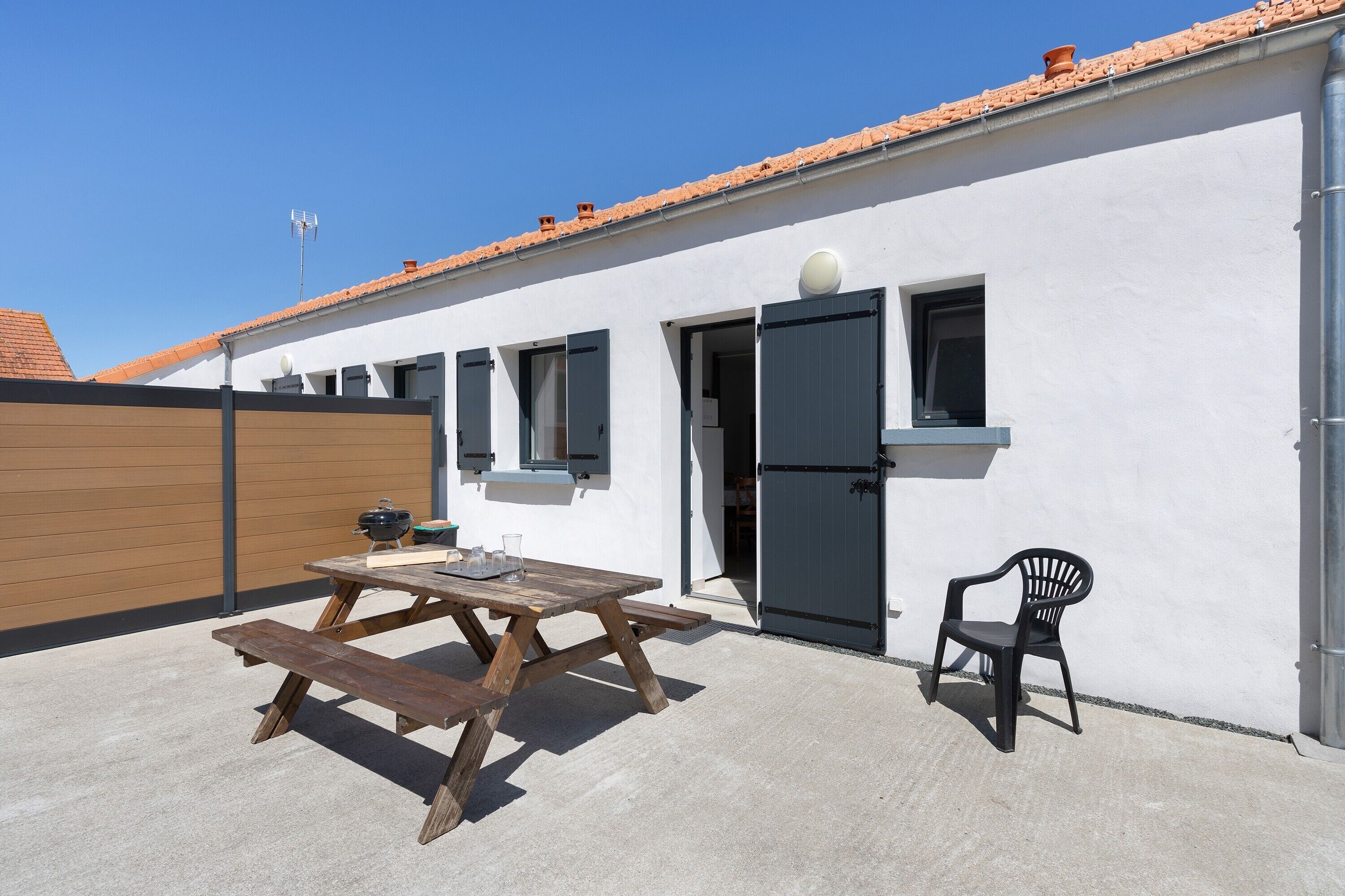 Een terras met een houten tafel en stoelen, een barbecue en een wit gebouw met een betegeld dak.