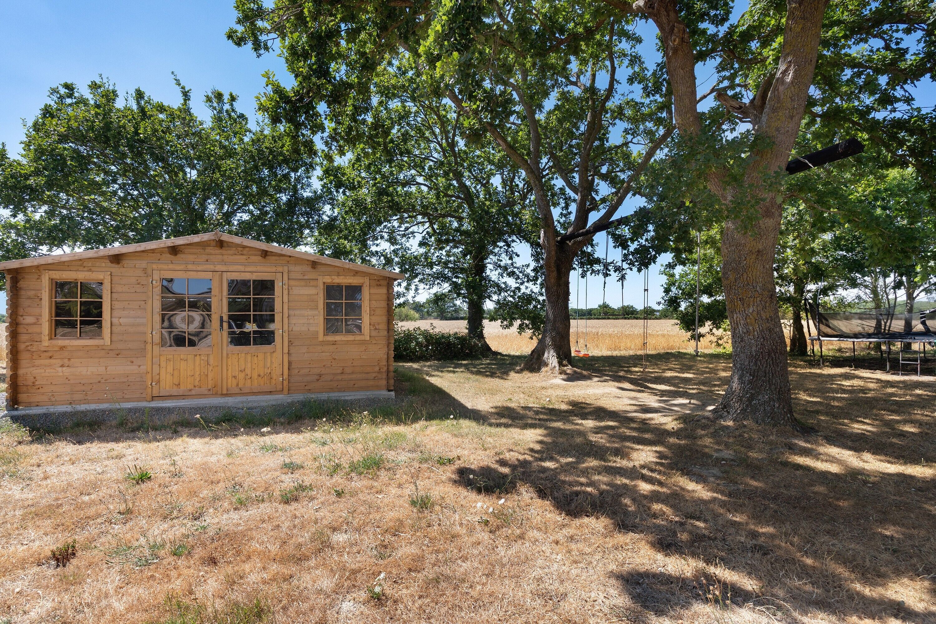Une cabane en bois avec un toit à pignon, de grandes fenêtres et une porte, entourée d’arbres et d’herbe sèche.