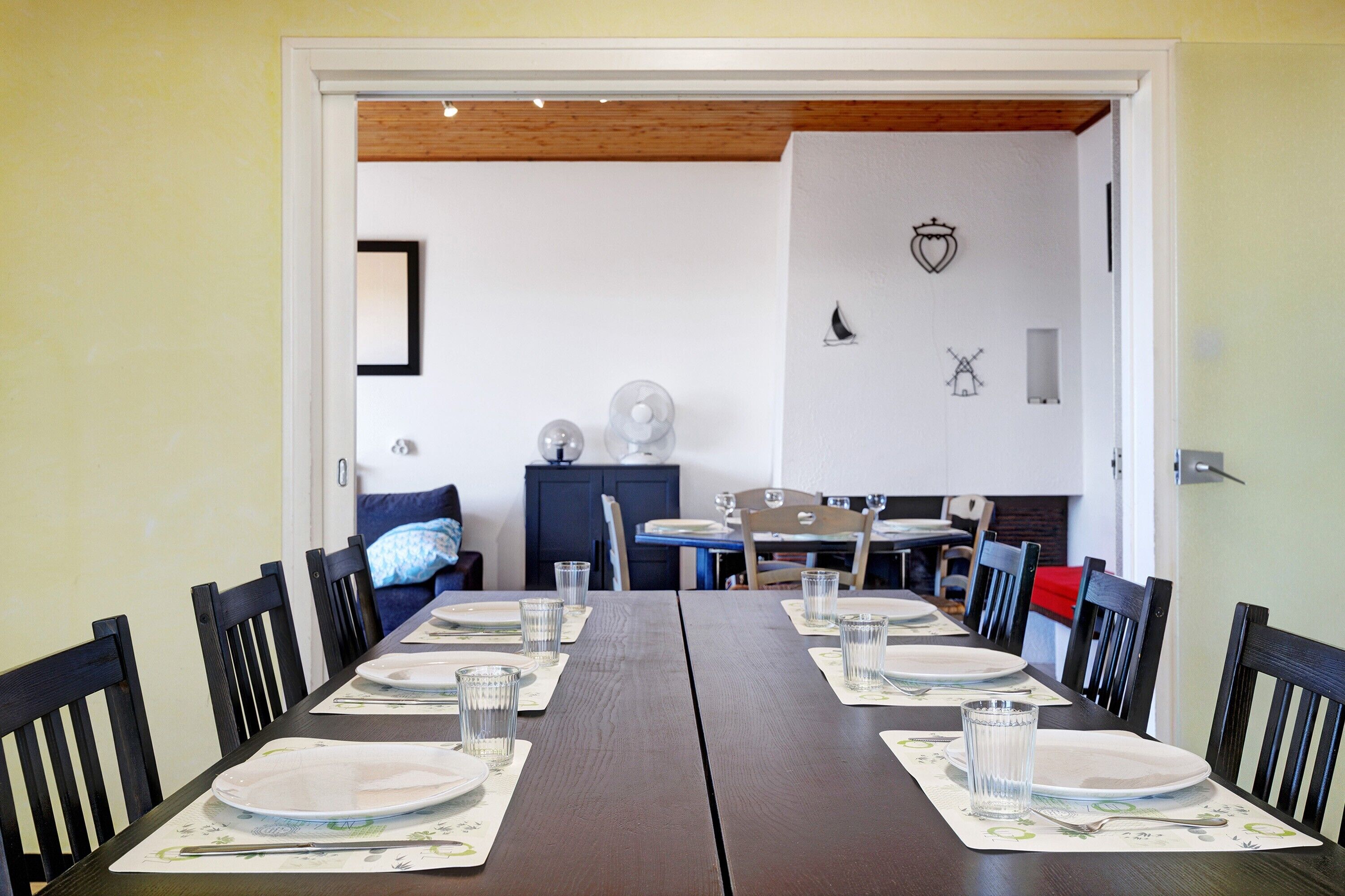 Un coin repas avec une longue table dressée pour deux personnes, des chaises et une vue sur une autre pièce où se trouvent un canapé bleu et un meuble.