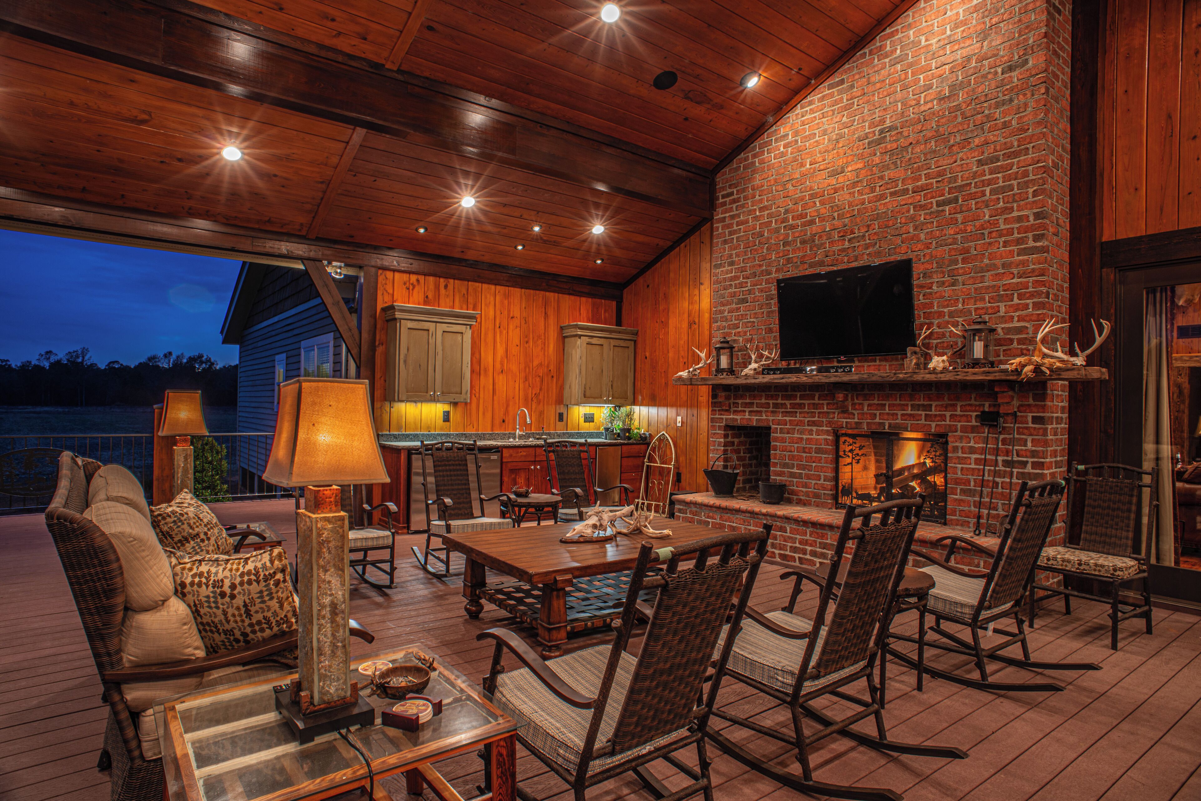 An outdoor patio with a brick fireplace, wooden furniture, and a television mounted on the wall.