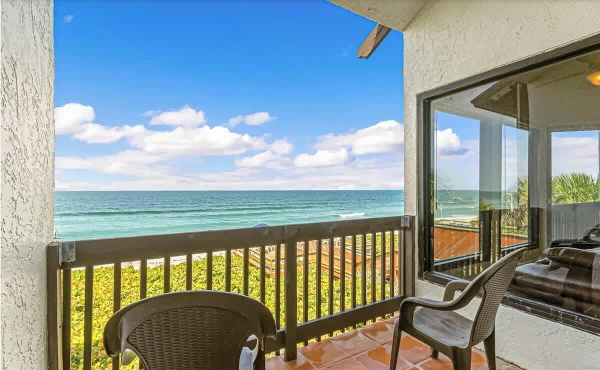 Ein Balkon mit Meerblick, ein Korbstuhl und eine Glastür zu einem Innenraum.