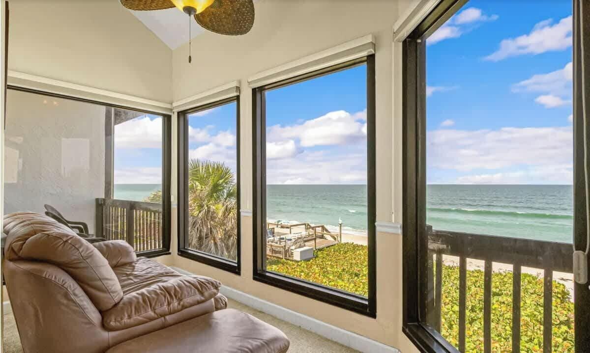 A room with a large window overlooking the ocean, a brown leather sofa, and a ceiling fan.
