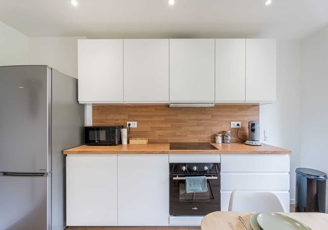 Una cocina compacta con gabinetes blancos, un refrigerador de acero inoxidable, horno integrado y cafetera sobre la encimera.