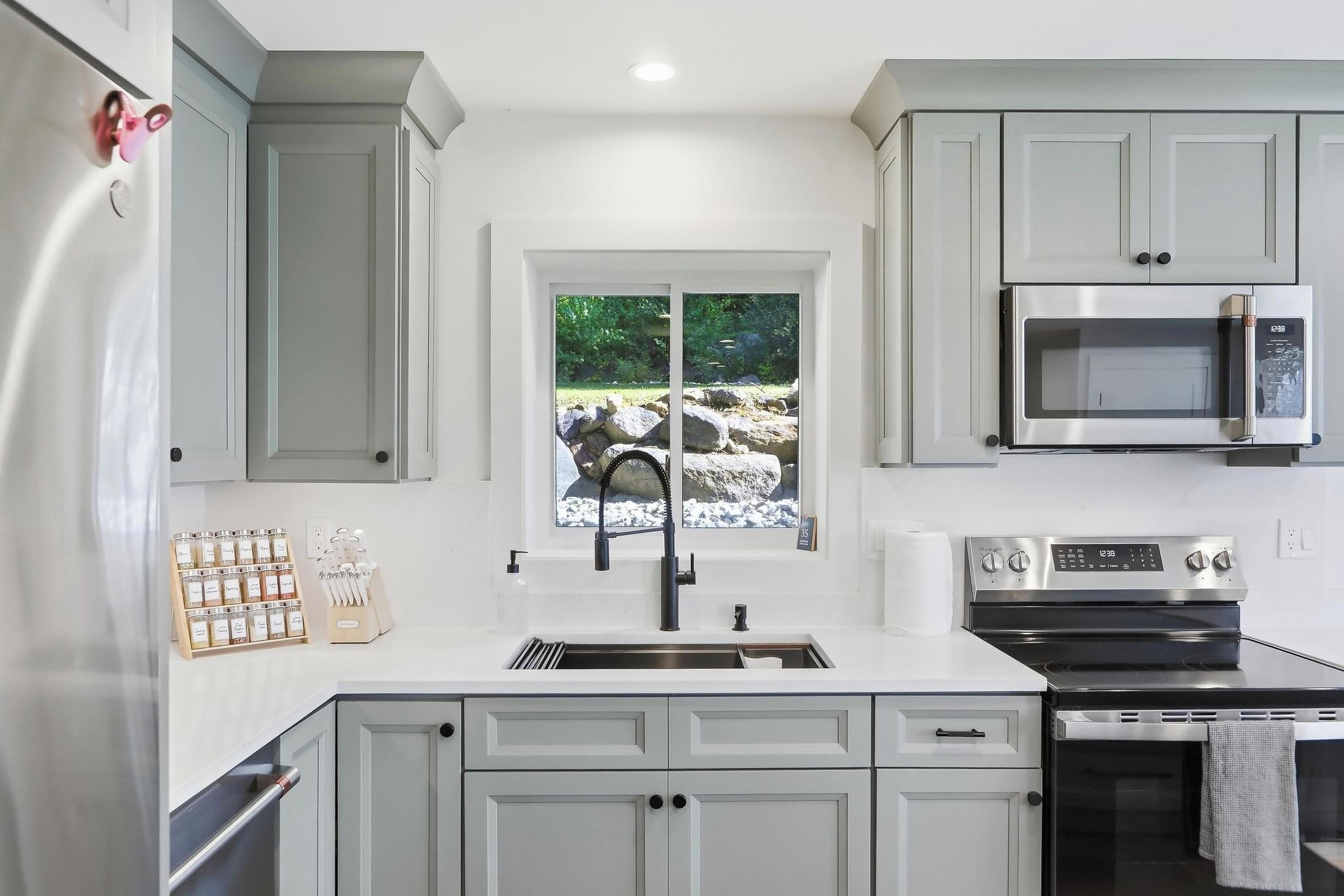 A modern kitchen with grey cabinets, stainless steel appliances, a window with a view of rocks and greenery, and a knife block on the counter.