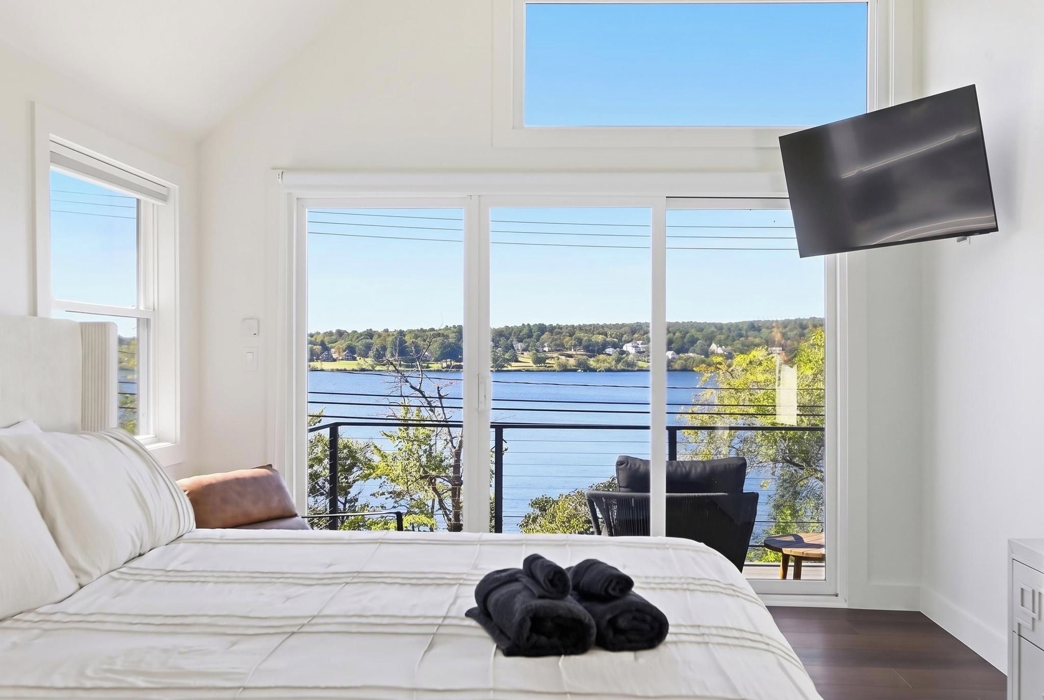 Ein Schlafzimmer mit einem großen Bett, einem an der Wand befestigten Fernseher und einem Balkon mit Blick auf einen See und Bäume.
