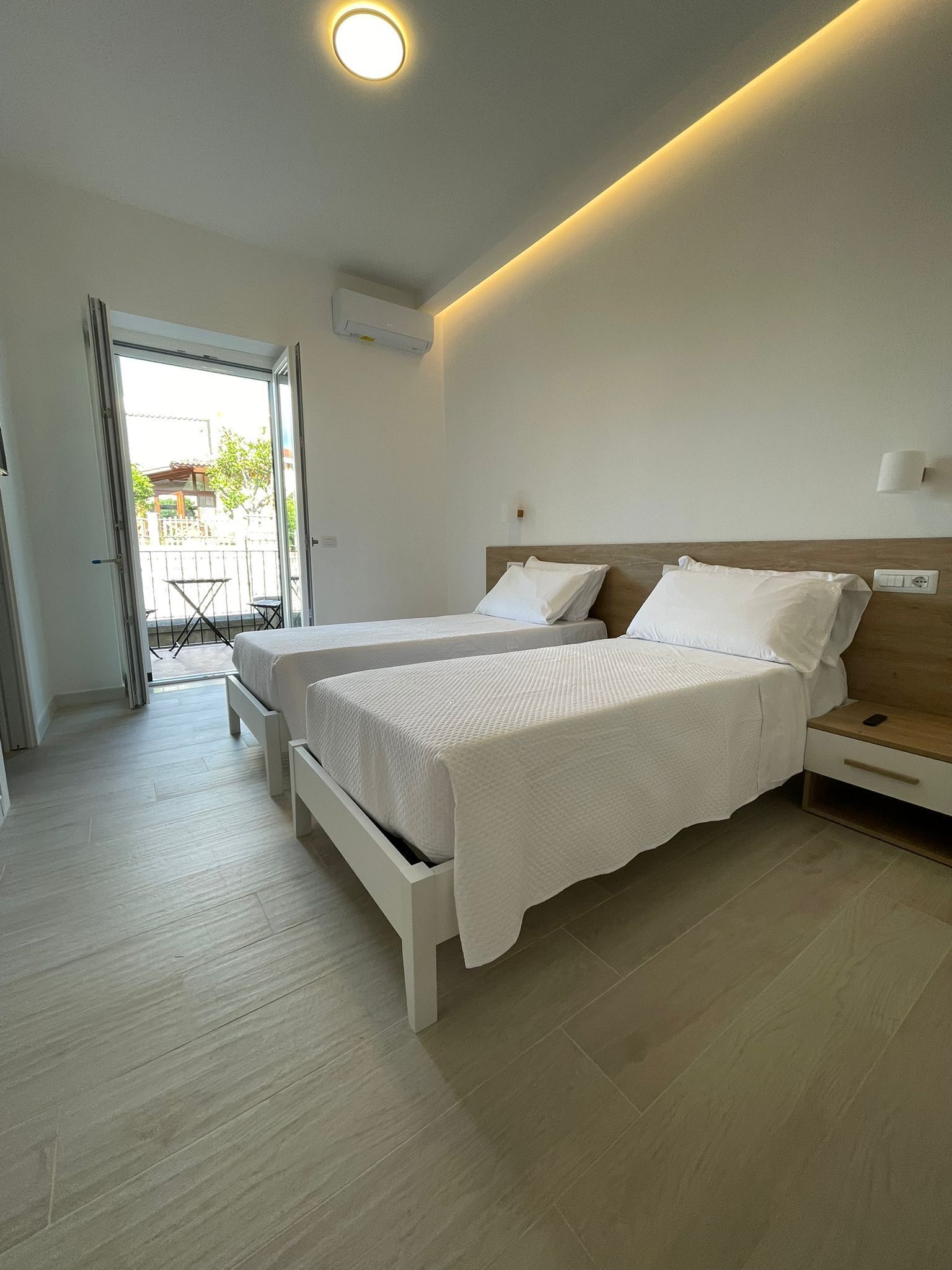 A hotel room with two beds, a wooden nightstand, a wall-mounted air conditioner, and a sliding glass door leading to an outdoor area.