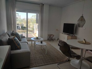Ein modernes Wohnzimmer mit Sofa, Couchtisch, Fernseher und einem Balkon mit Gartenmöbeln.