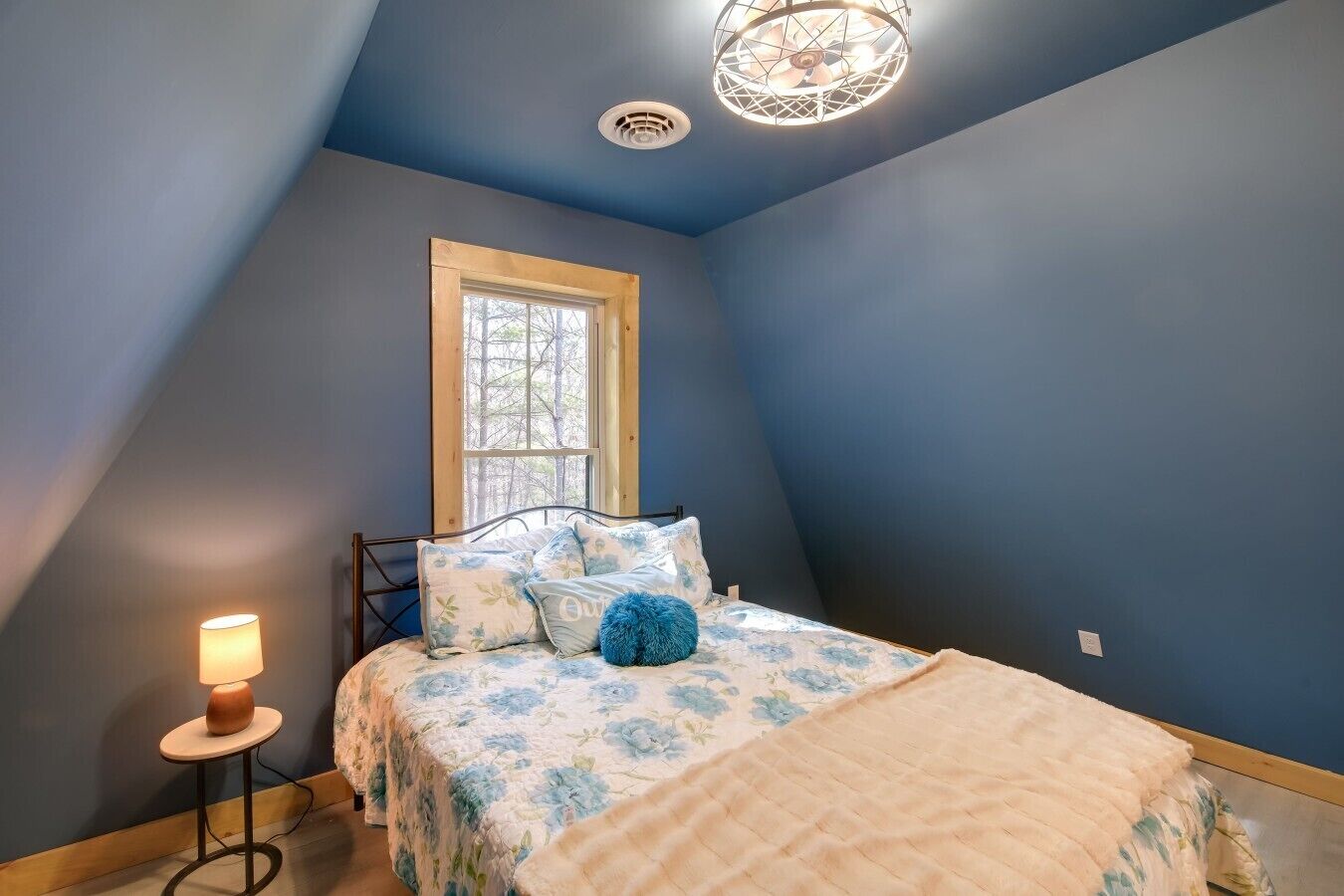 Ein Schlafzimmer mit geneigter Decke, einem Fenster, einem Bett mit Blumenmuster und einer runden Lampe sowie einem Deckenventilator.