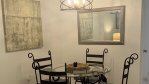 Uma sala de jantar com mesa de vidro redonda, quatro cadeiras de metal preto com assentos acolchoados, um lustre com três lâmpadas, um grande quadro abstrato e um espelho na parede.