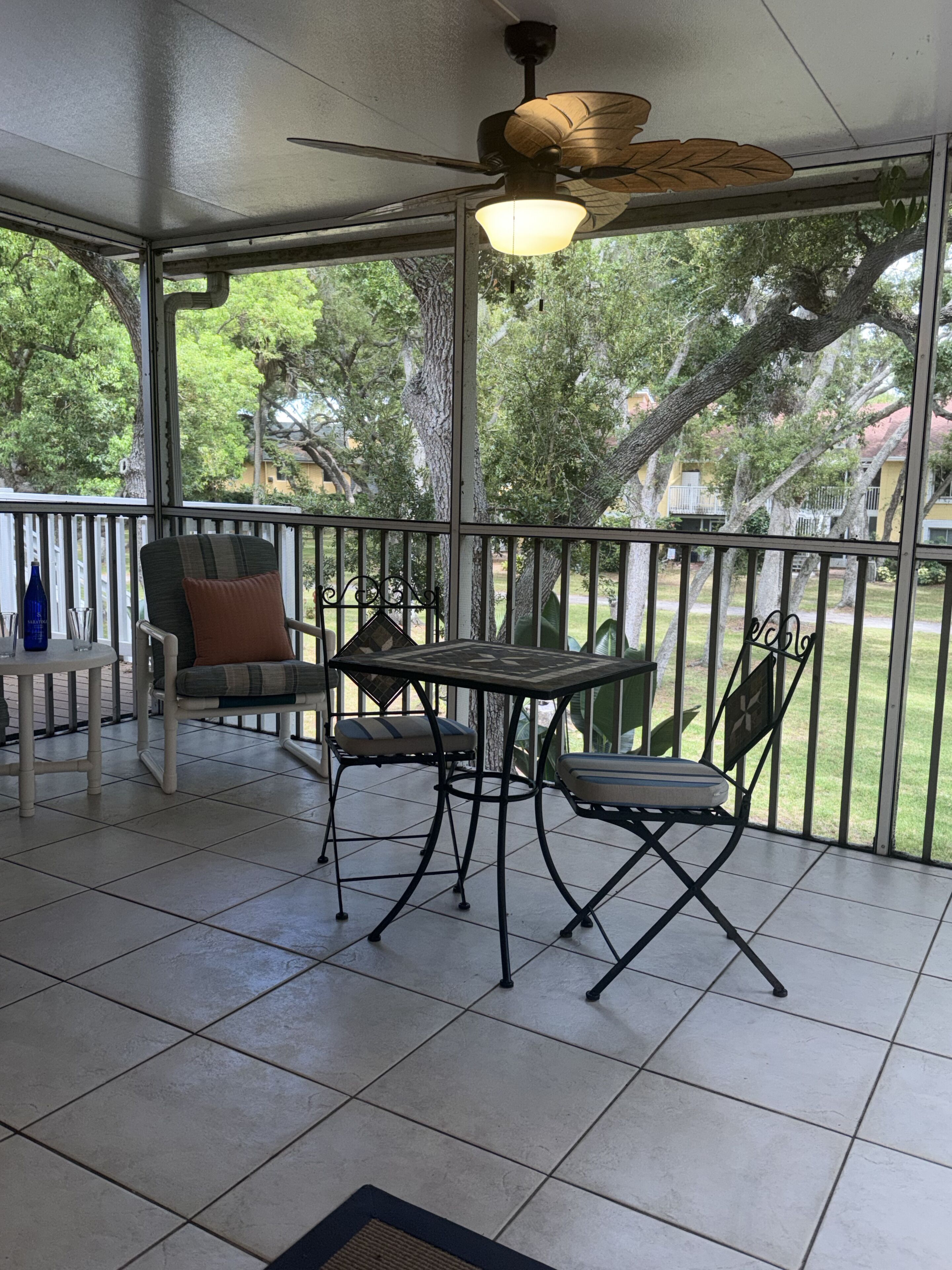 A covered patio area with a table and chairs, a ceiling fan, and a view of trees and buildings.