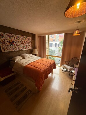 A bedroom with a bed, a chair, a window, and a wall with a colorful painting. - Beautiful Condesa Private Apartment (Ciudad de México)