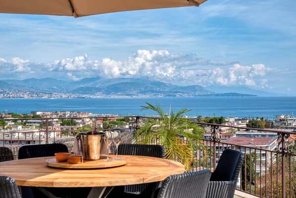A terrace with a wooden table, chairs, and a view of the sea and cityscape.