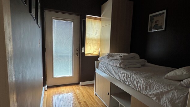 A bedroom with a wooden floor, a bed with a white mattress and pillows, a bedside table, a lamp, a wardrobe, and a door with blinds. - Contemporary 3-Bedroom Rowhouse on Capitol Hill (Washington)
