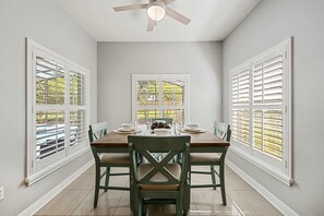 A dining area with a table set for four, surrounded by chairs, with a ceiling fan above and windows with blinds on the sides.