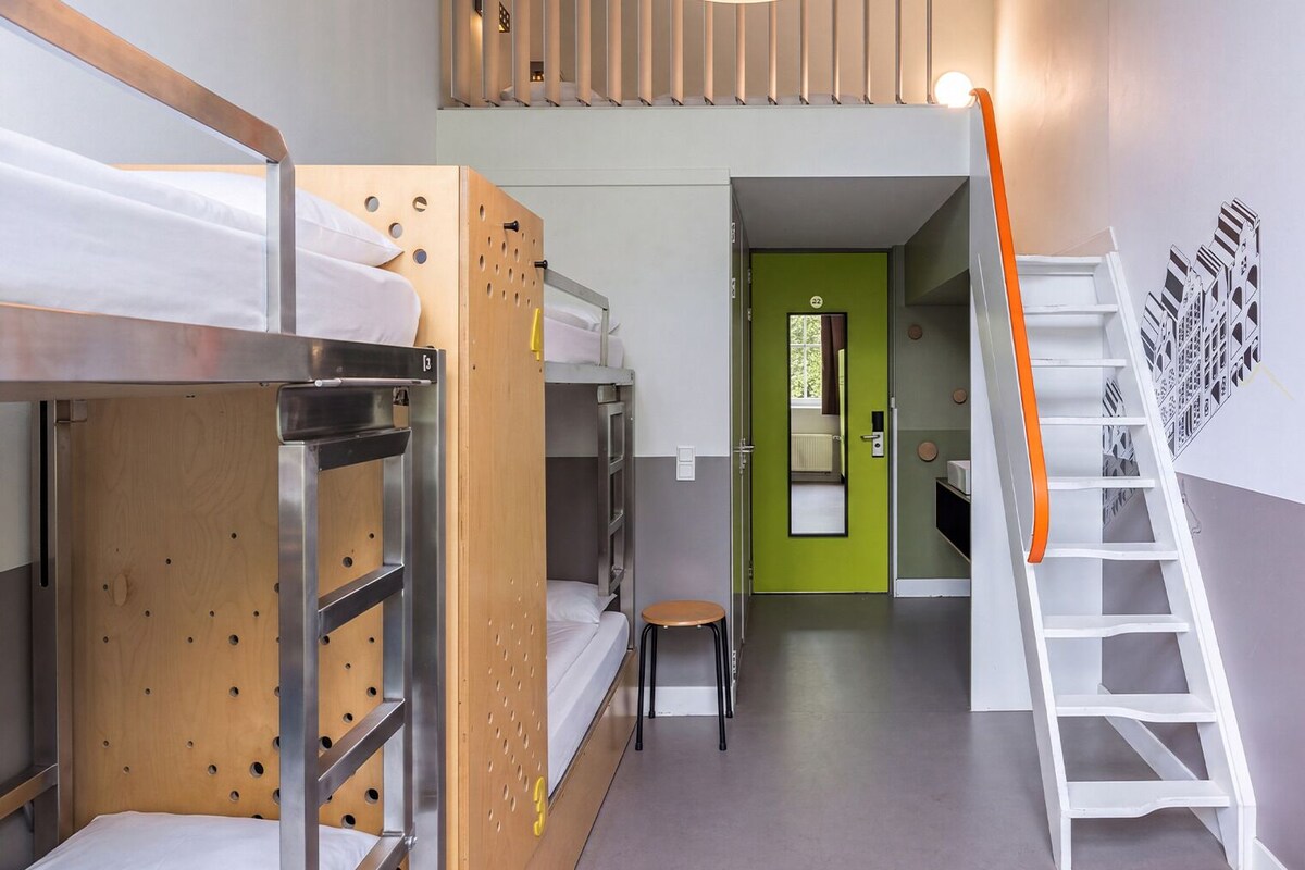 A dormitory room with bunk beds, a small stool, a green door, and a white staircase.