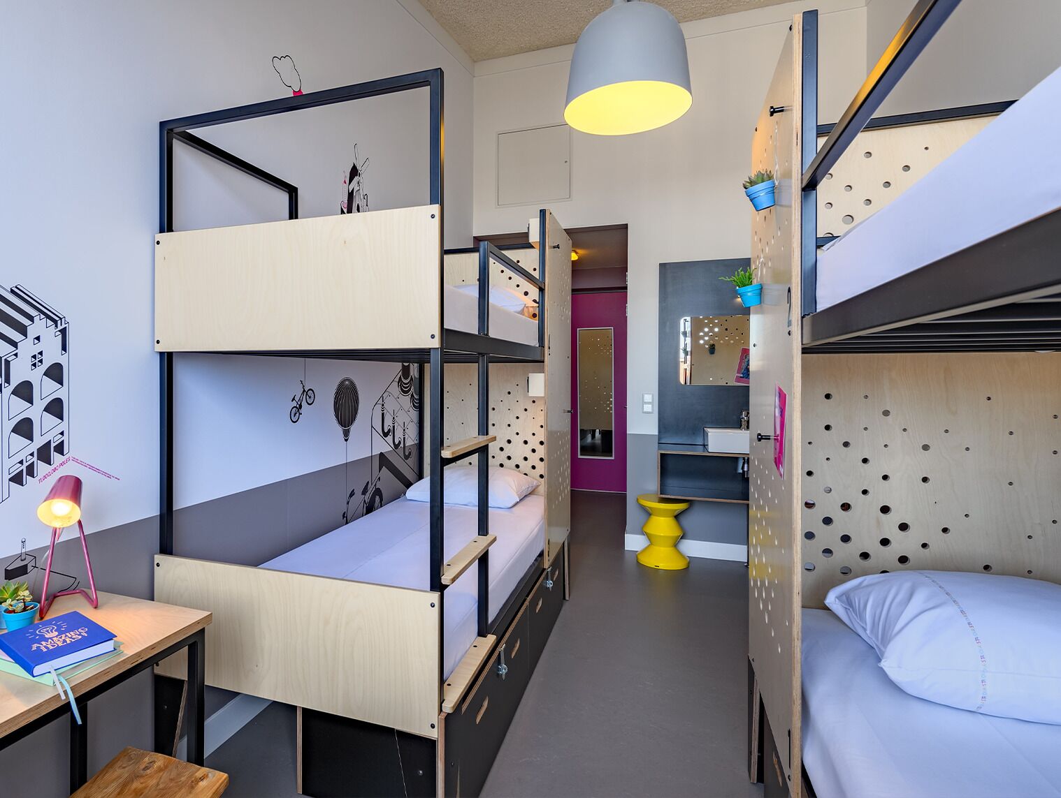 A dormitory room with bunk beds, a desk with a lamp, a small table, and a bathroom with a mirror and sink.