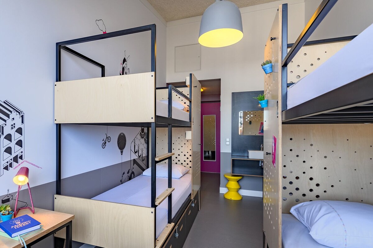 A dormitory room with bunk beds, a desk with a lamp, a small table, and a bathroom with a mirror and sink.