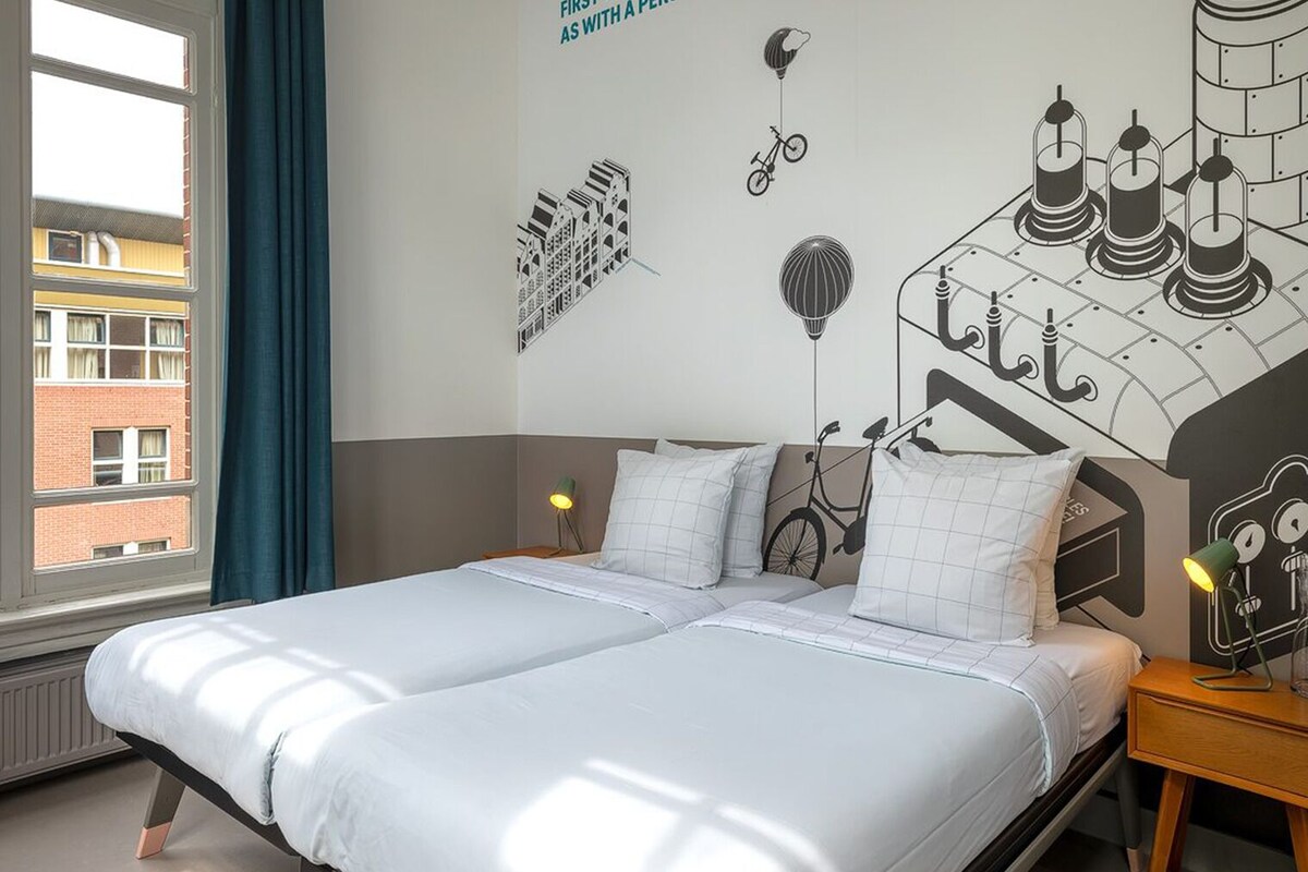 A double bed with white bedding, a nightstand with a lamp, and a wall mural with bicycles and hot air balloons.
