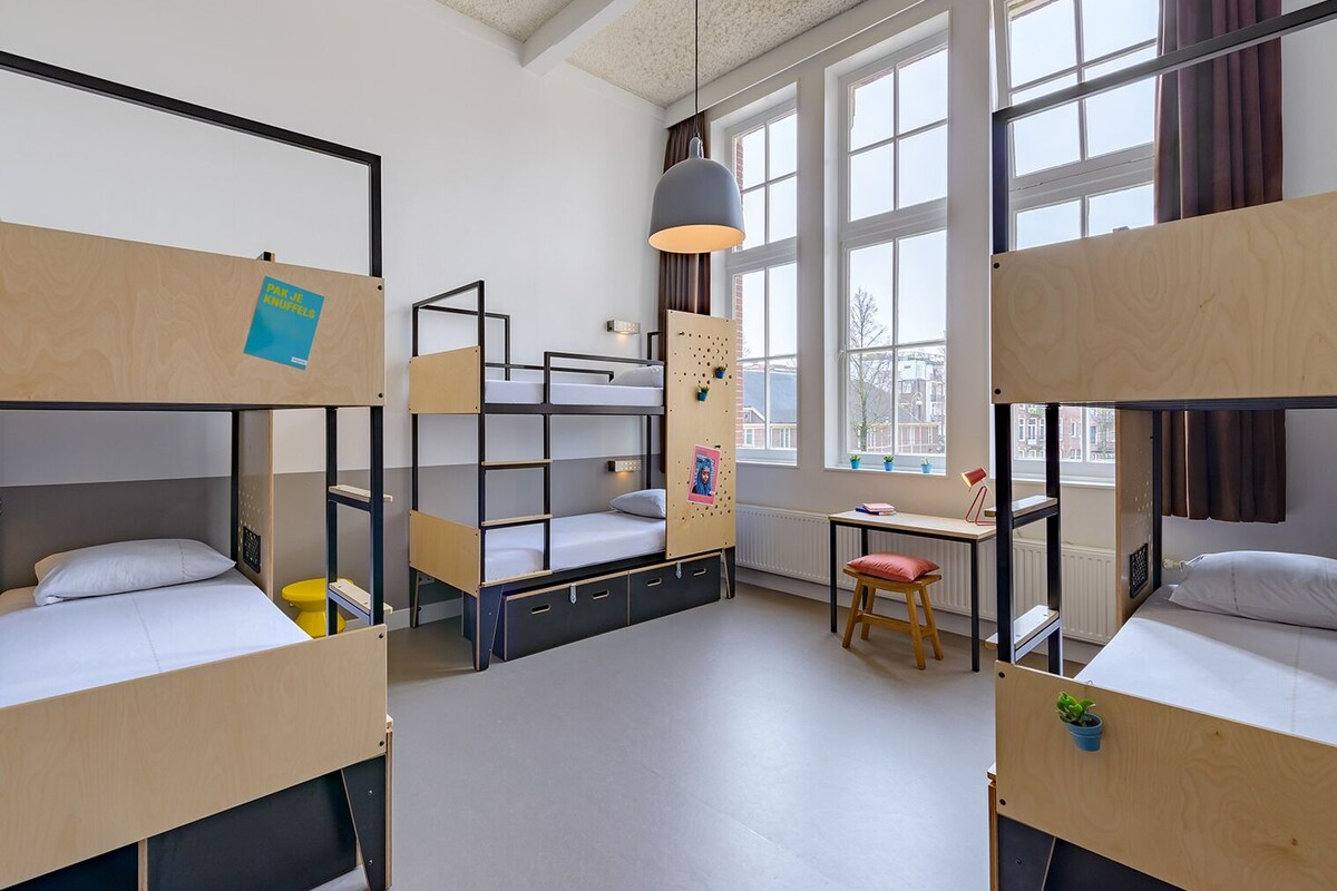 A dormitory room with bunk beds, a desk, a chair, and a hanging light fixture.