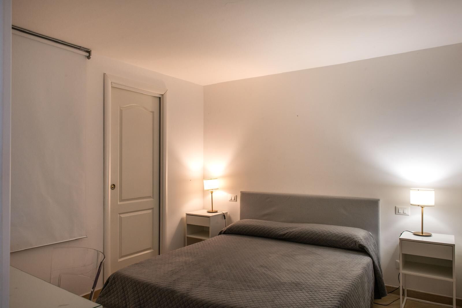 A simple, well-lit bedroom with a bed, two bedside tables with lamps, and a door.