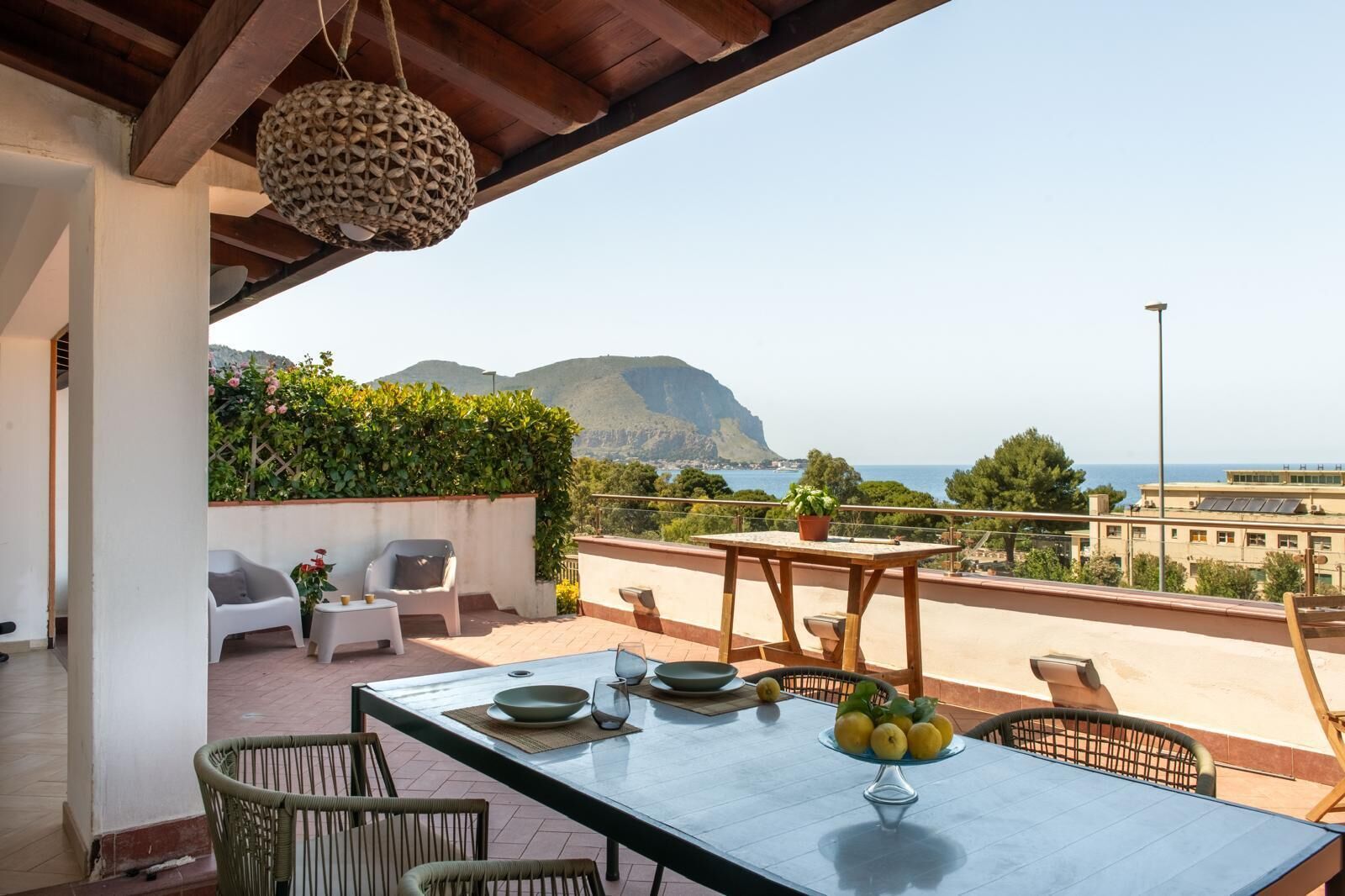 Une terrasse avec une table dressée pour le repas, des chaises, un luminaire suspendu et une vue sur la mer et les montagnes.