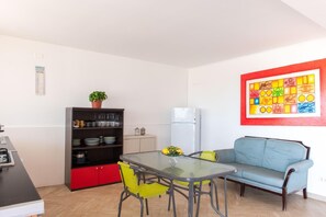 A compact living space with a kitchenette, a dining area with a glass table and chairs, a sofa, and a colorful abstract painting on the wall. - Mondello Sea View (Palermo)