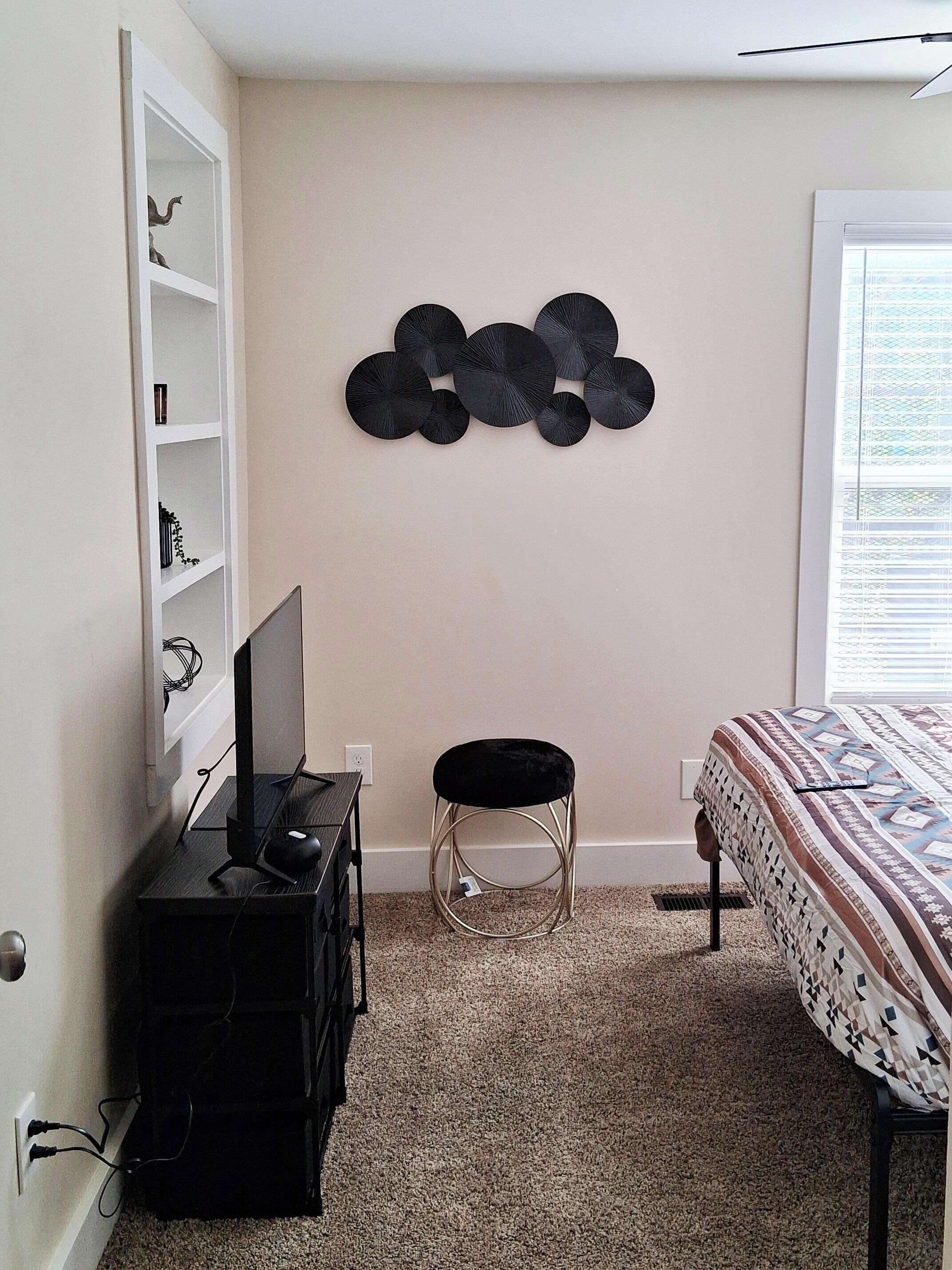 A bedroom with a bed, a desk with a computer, a chair, a wall-mounted shelf, and a decorative wall piece.