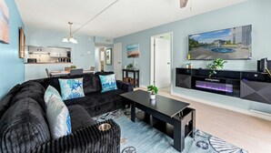 Un salon moderne avec un canapé, une table basse et un téléviseur à écran plat. On aperçoit une cuisine à l’arrière, avec des placards et un coin repas.