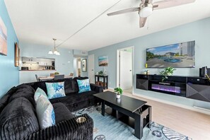 Ein modernes Wohnzimmer mit einer Couch, einem Couchtisch und einem Flachbildfernseher. Im Hintergrund befindet sich eine Küche mit Schränken und einem Essbereich.