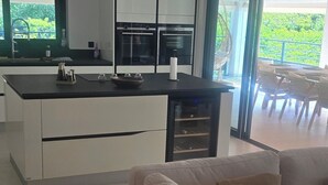 En moderne stue med en beige sofa, et køkken med hvide skabe og et spiseområde med fletmøbler.