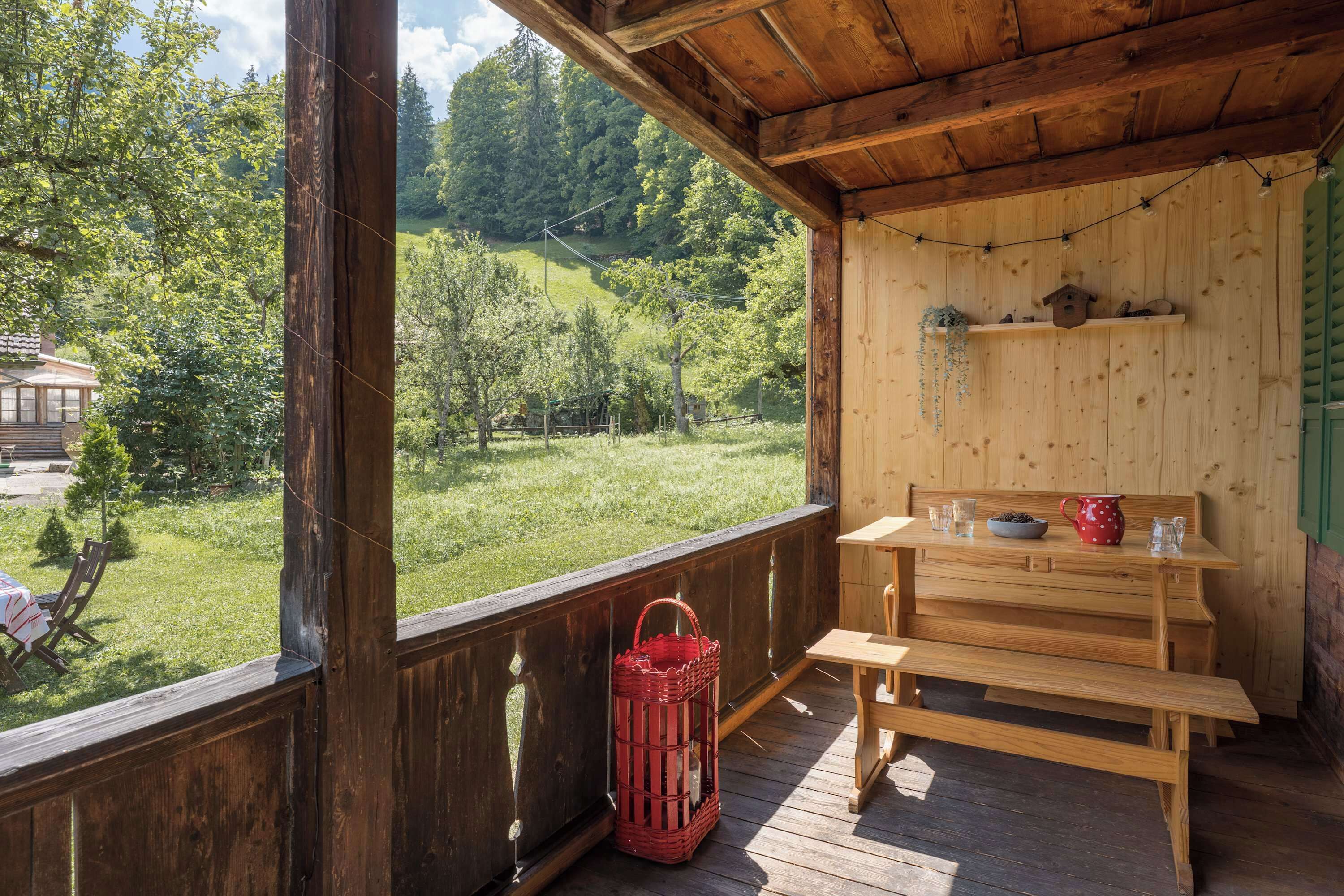 Une terrasse en bois avec un banc et une table, donnant sur un paysage verdoyant avec des arbres et une maison.